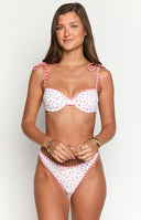 Model is wearing a white bikini set featuring a ditsy red floral print. The bikini top has underwire cups and is trimmed with red and white gingham fabric- which also forms the tie-shoulder straps