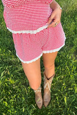 Top-down view of red gingham shorts with white broderie lace trim on the hem and waist