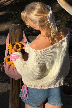Rear view of a cream chunky knit sweater with pink lacing detail on the off-shoulder neckline, sleeves, and hem