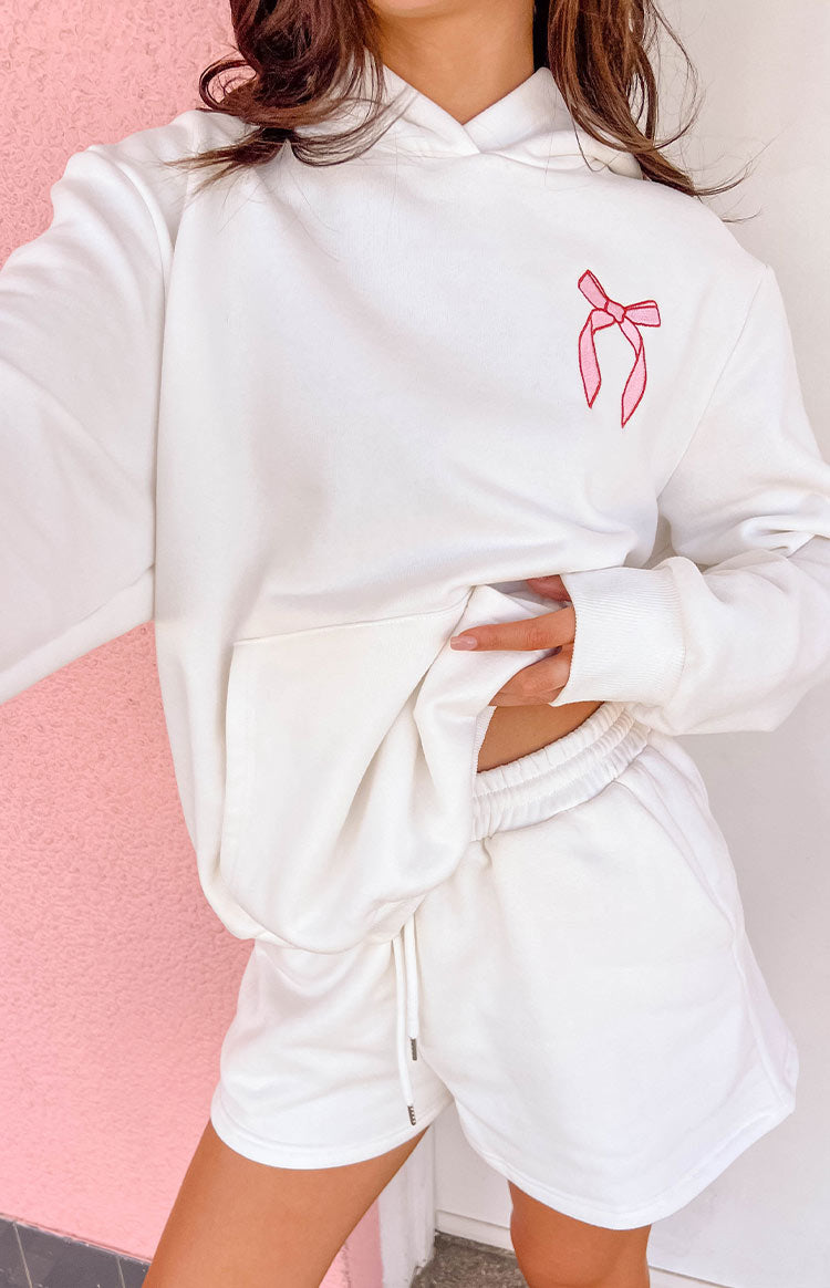 White hoodie with a pink embroidered bow on the chest, a large front kangaroo pocket, and ribbed cuffs