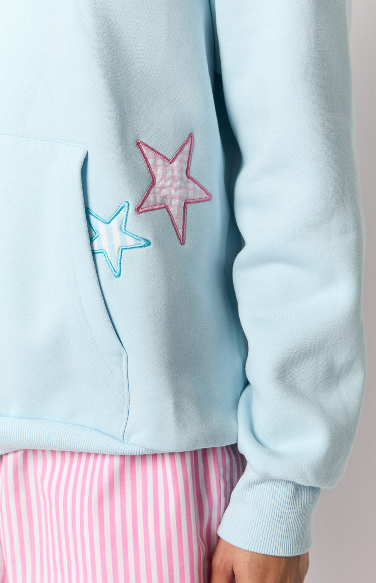 Close-up of light blue hoodie featuring kangaroo pocket with pink gingham and blue striped star patches, ribbed cuff