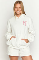 Brunette girl wearing a big oversized white hoodie with pink bow designs embroidered on the chest and sleeves. Girl is wearing white ruffled shorts.