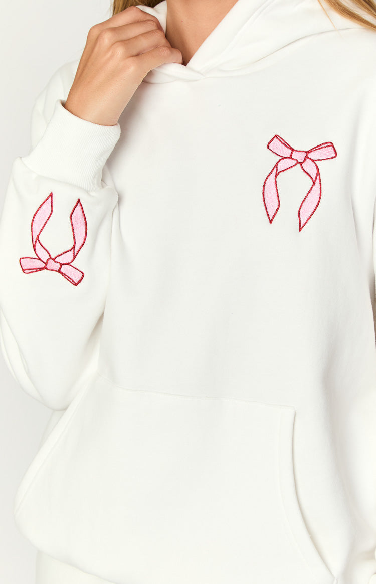Close-up of a white hoodie showing pink bow embroidery on the chest and left sleeve cuff, with a ribbed cuff and kangaroo poc