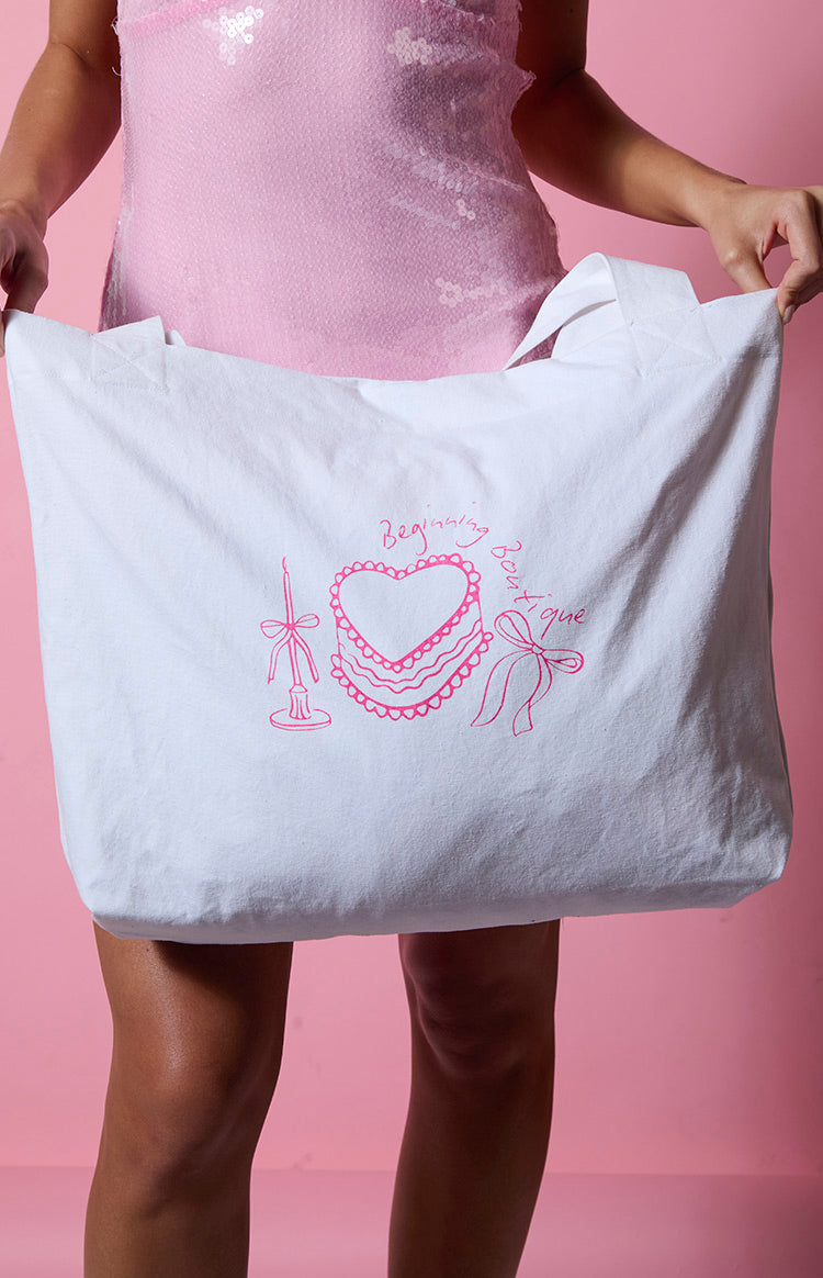 White canvas tote bag with a pink graphic of a heart-shaped cake, candle, and bow illustration