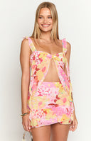 Girl is wearing a chiffon ruffled top with a bright floral print with pinks, yellows, and reds. She is wearing the matching skirt and holding a woven handbag with gold jewellery.