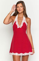 a blonde model wears a red polka dot mini dress with white lace detailing and a gold bracelet with red rosed in the background