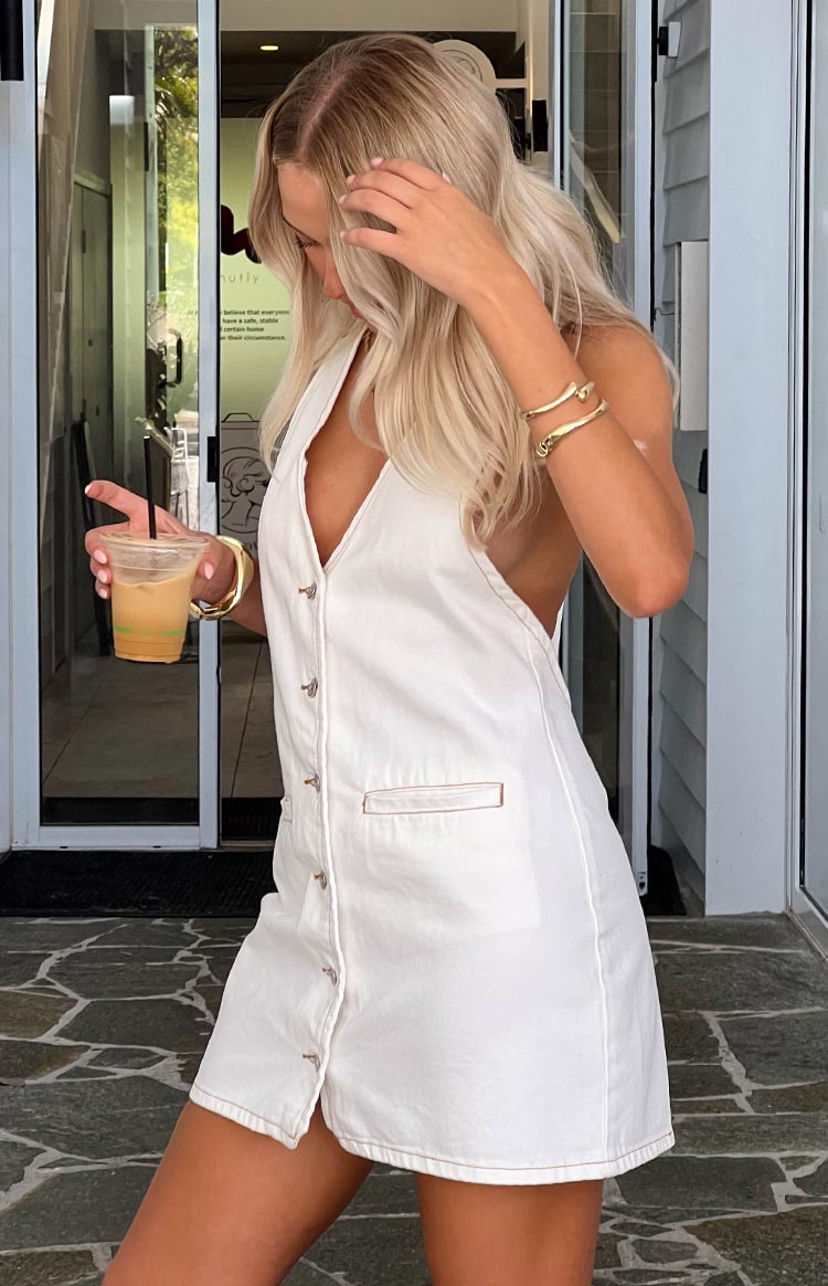 Side view of a white denim halter neck mini dress with a deep V-neck, front button closure, and contrast stitching