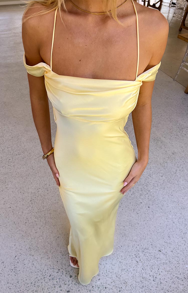 A close-up of a pale yellow satin formal maxi dress with an elegant off-the-shoulder draped neckline and delicate spaghetti straps- highlighting its refined design.