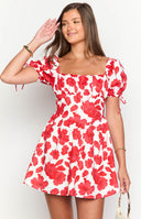 Brunette girl wearing a red and white floral mini dress with gold jewellery and holding a drink.