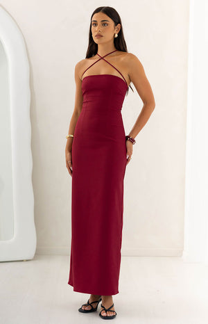 Our model wearing a form-fitting floor-length maxi dress in a burgundy color. This maxi features a strapless contoured bodice and thin halter straps that crisscross across the chest and wrap around the neck. The skirt has a straight column silhouette.