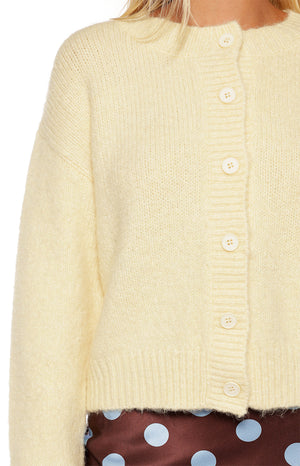 A close-up of a fuzzy light yellow knit cardigan featuring a crew neckline and a full placket of white buttons.