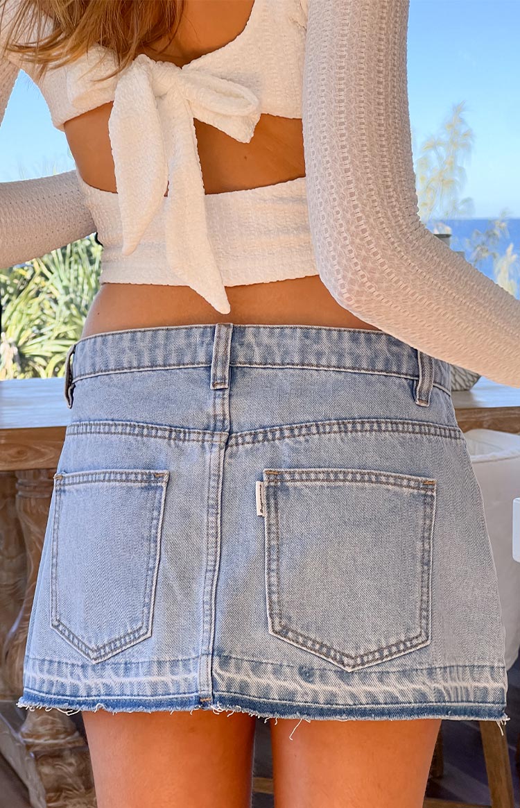 Back view of a light wash distressed denim mini skirt- paired with a white textured long-sleeve top with a large bow detail at the back.