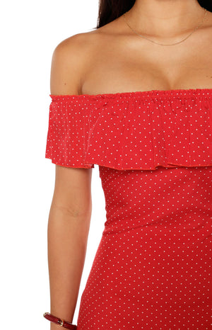 A close-up of a red off-the-shoulder mini-dress with a small white polka dot pattern highlighting the wide elasticized ruffle neckline and fitted bodice.