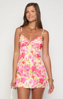 A model with blonde hair wears a floral mini dress in shades of pink, yellow, and white with a ruched bodice. They are wearing a gold star necklace and multiple gold bracelets and rings. The background shows a textured white wall and a light pink railing.