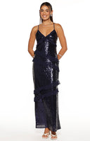 Our model wearing a floor-length maxi dress in a navy blue maxi dress. This fitted gown is completely covered in shimmering sequins and is detailed with horizontal cascading sheer fabric ruffles across the bodice and skirt. It features thin spaghetti straps and a V-neckline also trimmed with ruffles.