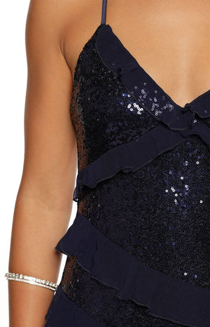A close-up view of our model wearing a navy blue sequined maxi dress. This maxi features a V-neckline and is completely covered in dark shimmering sequins. It is embellished with thin horizontal navy blue sheer ruffles across the bodice.