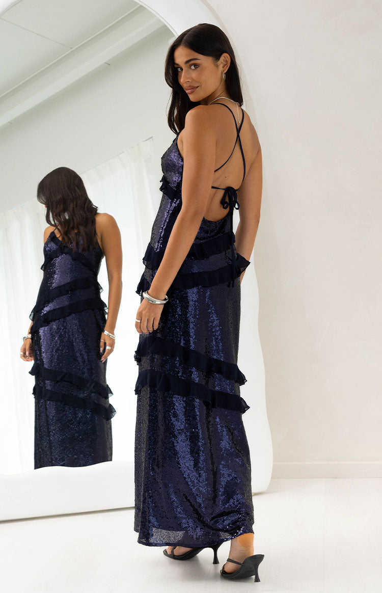 Rear-side view of a navy sequin maxi dress with criss-cross back straps, low tie detail, and tiered ruffles