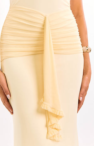 A close-up of our model's waist and hips showing a cream maxi dress with a ruched draped chiffon panel and a long ruffled cascade detail.