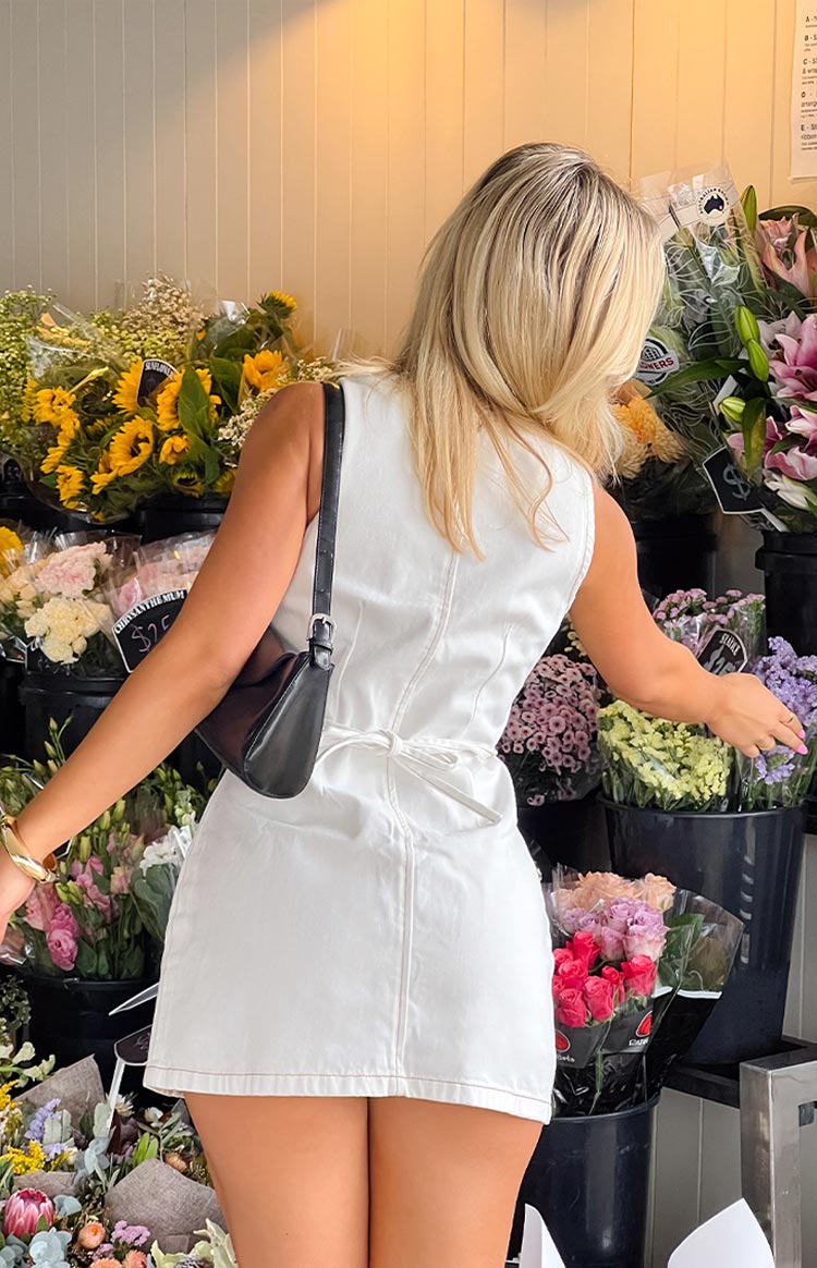 Back view of a crisp white wash denim mini dress- featuring a sleeveless design and a unique self-tie detail at the waist- offering a casual yet stylish silhouette.
