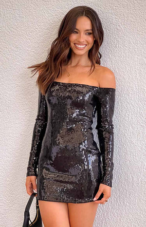 brunette girl wearing a black sequin long sleeve mini party dress with of shoulder neckline, styled with a gold necklace and black handbag