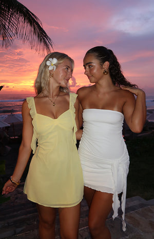Two girls in front of a purple and orange sunset. Girl wearing a yellow mini dress and gold jewellery and girl wearing a white strapless mini dress.