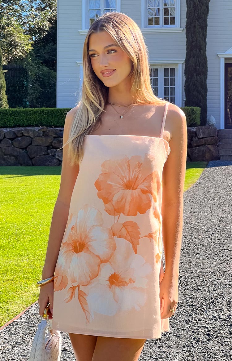 A light peach mini dress adorned with large- soft orange and white hibiscus floral prints- featuring a simple square neckline and delicate spaghetti straps- suitable for a casual summer event.