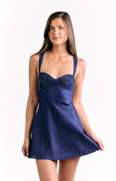Our model wears a short fit-and-flare slip dress in a deep satin navy blue fabric. The dress features thin spaghetti straps and a bodice with a sweetheart neckline. The top section of the bust is ruched and embellished with a subtle dark metallic sparkle. The skirt is smooth flowing and short.