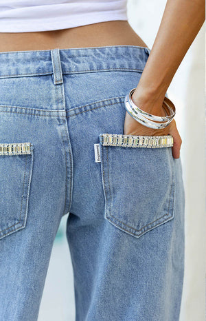 A close-up view of the back of a pair of light-wash denim jeans. The standout feature is the crystal embellishment—two horizontal rows of rectangular clear baguette-cut rhinestones are set along the top edge of each back patch pocket. The jeans have a straight or wide-leg cut and belt loops.
