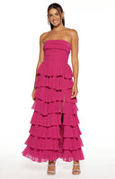Our model wearing a dramatic strapless maxi dress in a vibrant fuchsia color. This maxi features a fitted straight bodice and a voluminous skirt composed of multiple horizontal tiers of sheer ruffles. The skirt has a high slit to the thigh revealing her legs and a pair of beige block-heeled sandals with rhinestone straps. Thin lace-up ties are visible on the back/side of the bodice.