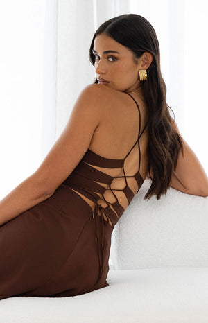 A back view of our model highlighting the intricate back detail of a brown maxi dress. This maxi features an open multi-strap back with horizontal strips of fabric laced up in a corset style with thin vertical ties.