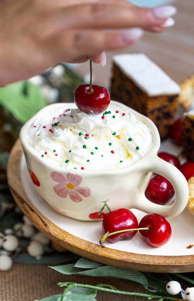 Close-up of a cream ceramic mug with pink floral and red cherry print, filled with whipped cream, sprinkles, and a single cherry