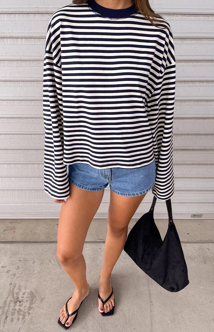 Navy blue and white horizontal striped long-sleeve top with a mock neck, paired with blue denim shorts. Black square-toe thong sandals and a black shoulder bag are accessorized.