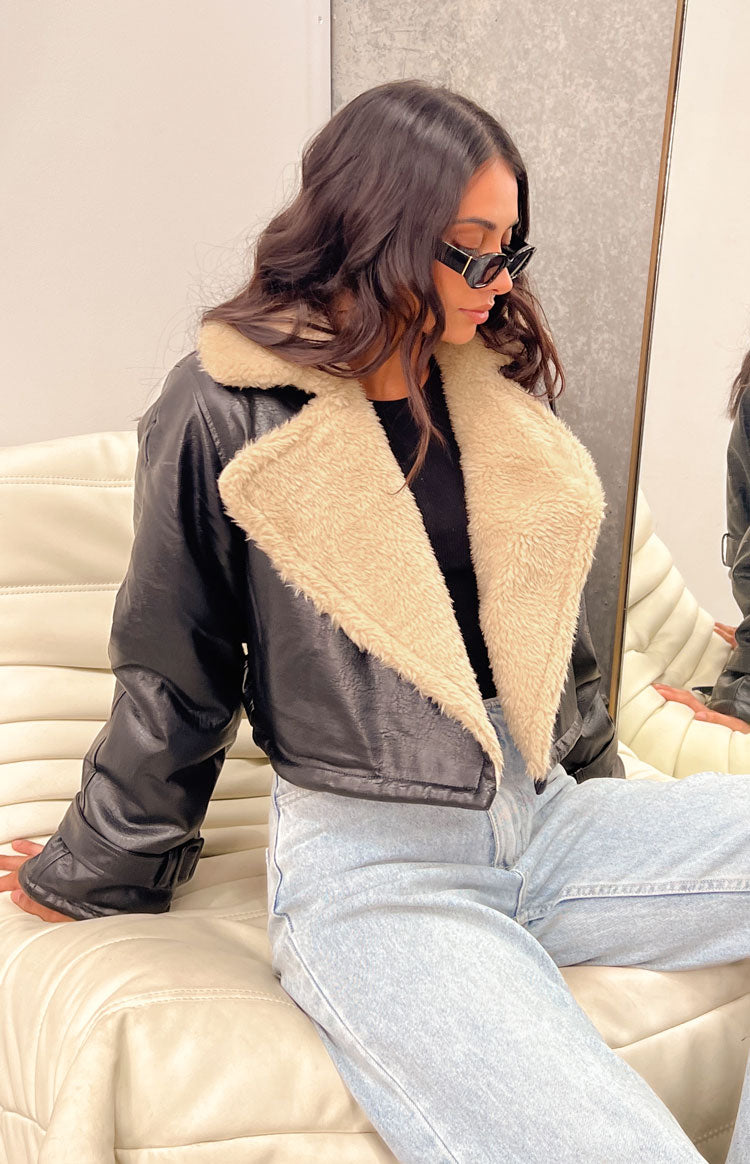Side view of a black faux leather cropped jacket with a wide cream shearling collar and buckled cuffs