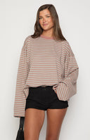 A person with long blonde hair wears a horizontally striped long-sleeved shirt in shades of pink and beige, paired with black textured shorts. They are standing against a white brick wall.