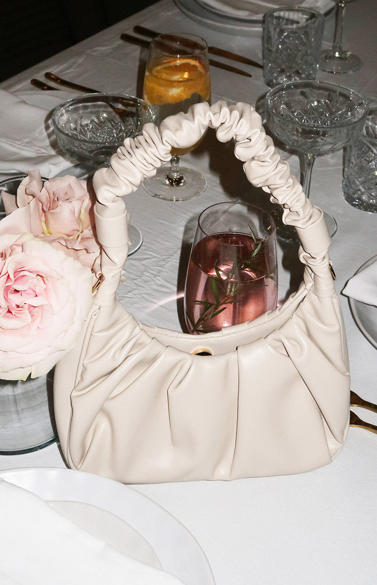 Cream handbag with a ruched top handle, soft pleated body, and a gold magnetic snap closure