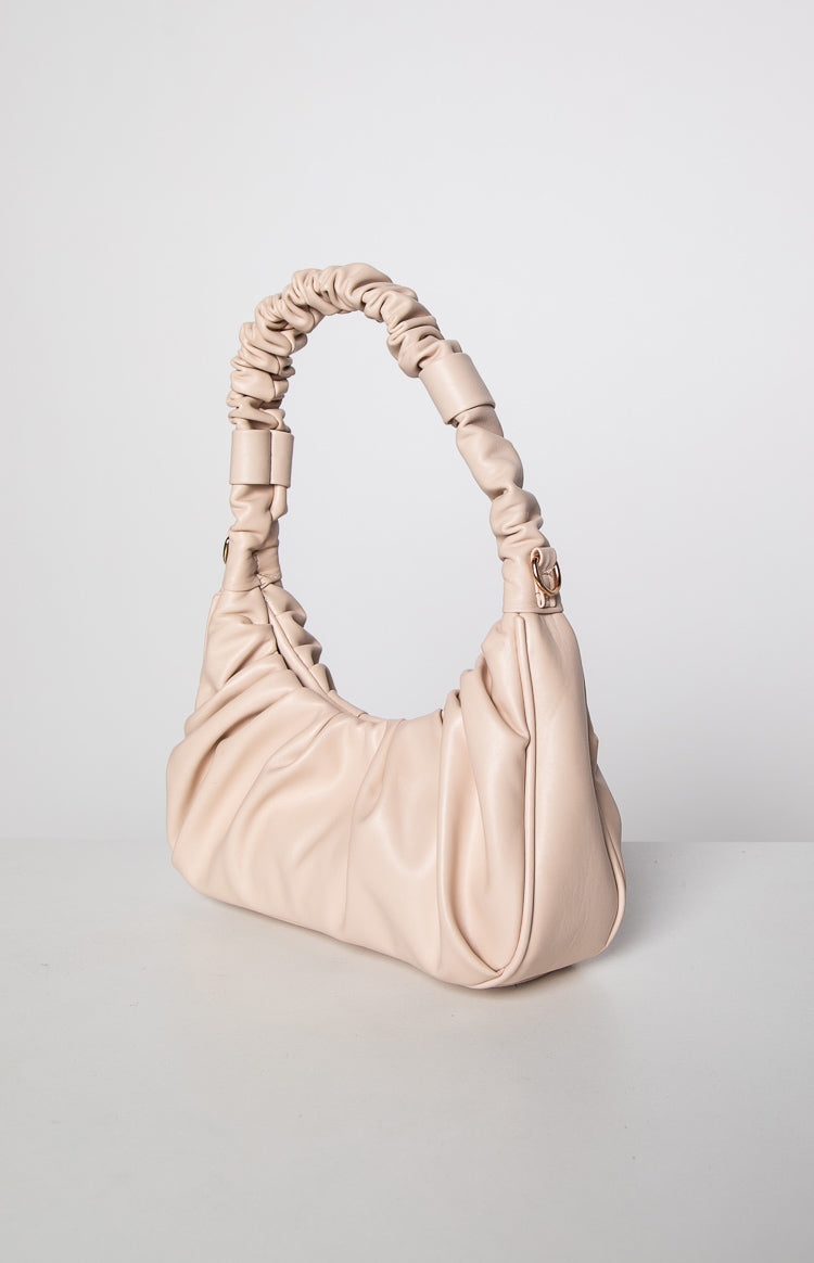 Side view of a cream ruched faux leather handbag with a scrunchie-style handle and gathered body