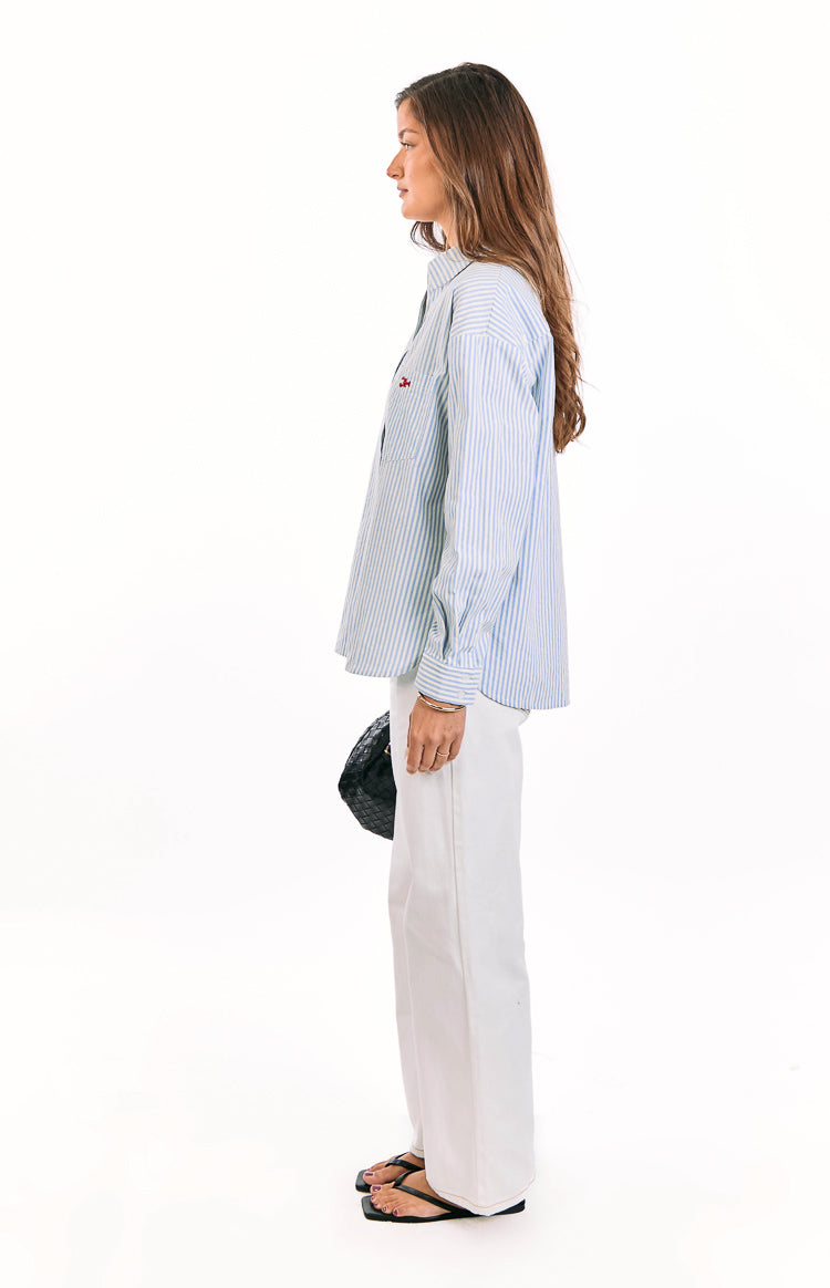 Side view of a light blue and white striped button up shirt with a red embroidered detail on the chest pocket