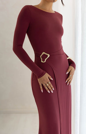 Our model wearing a form-fitting long-sleeve maxi dress in a deep burgundy features of a smooth stretch fabric with a modest boat neckline. This maxi has a faux wrap design at the waist with a large asymmetric gold buckle accent. The skirt features a wrap slit overlay from the waist down.
