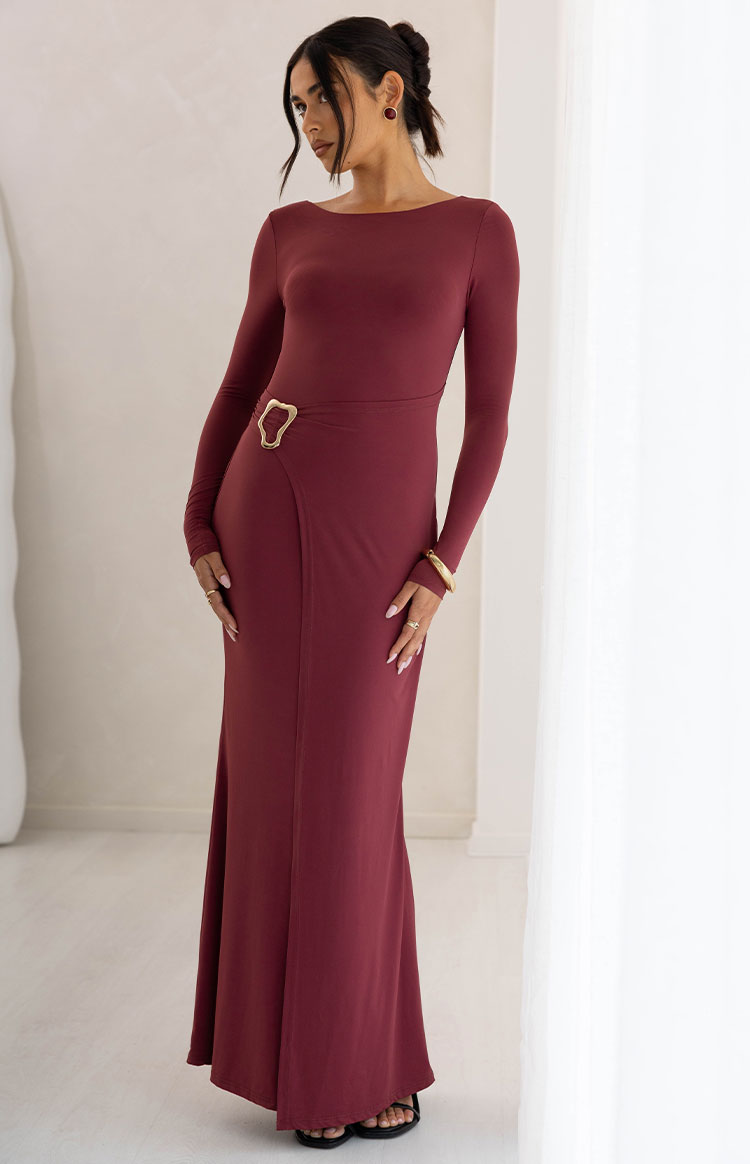 Burgundy long sleeve maxi dress with a boat neck and gold abstract buckle detail at the wrap waist