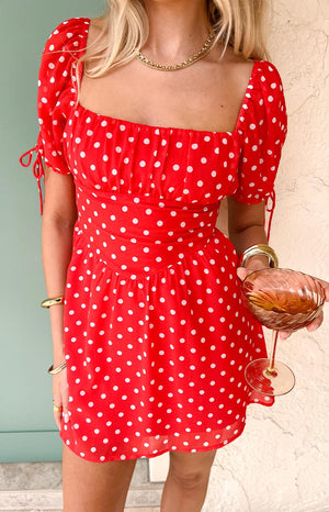 A person models a red mini dress with white polka dots and short puff sleeves. They are wearing a gold chain necklace and multiple gold bracelets and holding a stemmed glass with a brown beverage.
