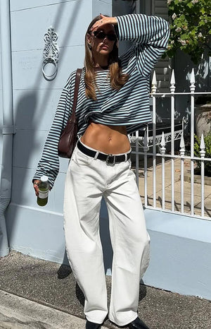 A person wearing a striped long-sleeved shirt, white pants with a black belt, and sunglasses stands outside a light blue building. They are holding a drink in one hand and have a brown bag slung over their shoulder.