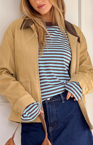 A person models a tan jacket with a brown corduroy collar over a blue and brown striped long-sleeved shirt and dark blue jeans. The person has long blonde hair and is wearing a brown shoulder bag.