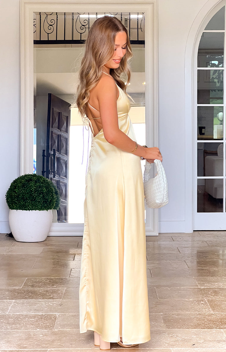 Side view of a pale yellow satin maxi dress with a low back, thin criss-cross lace-up straps, and ruched side detail
