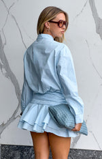 Rear view of a light blue long sleeve mini shirt dress with a cinched waist and tiered ruffled skirt
