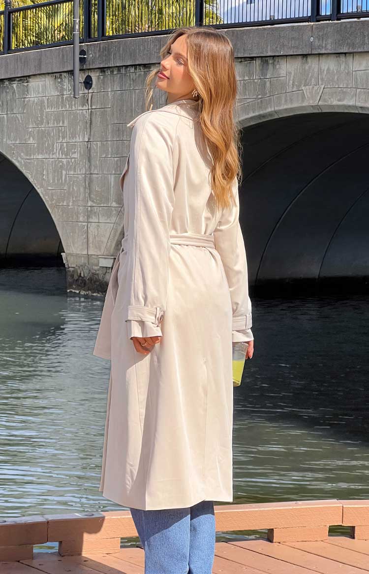 Rear-side view of a cream belted trench coat with tied waist and cuff straps