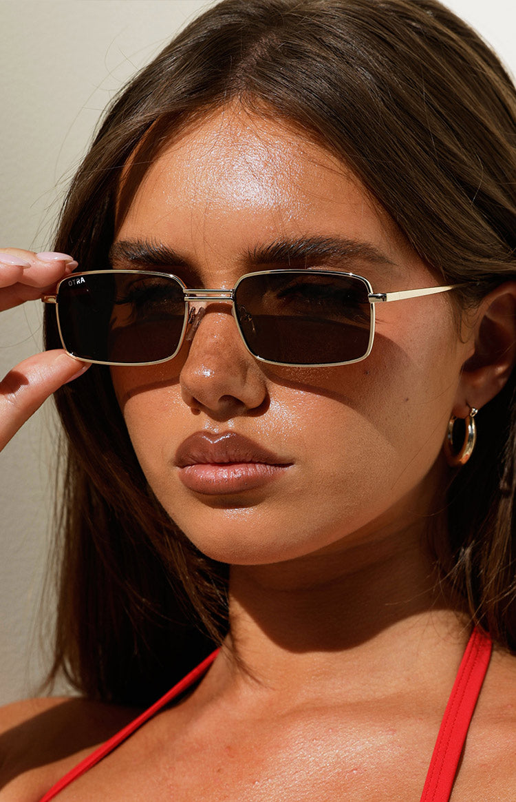 Close-up of gold metal square frame sunglasses with brown lenses, showing the OTRA logo on the left lens