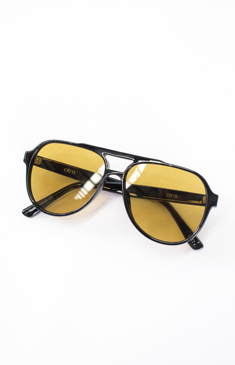 Angled view of black aviator sunglasses with yellow lenses, showing the otra logo and STEVIE text
