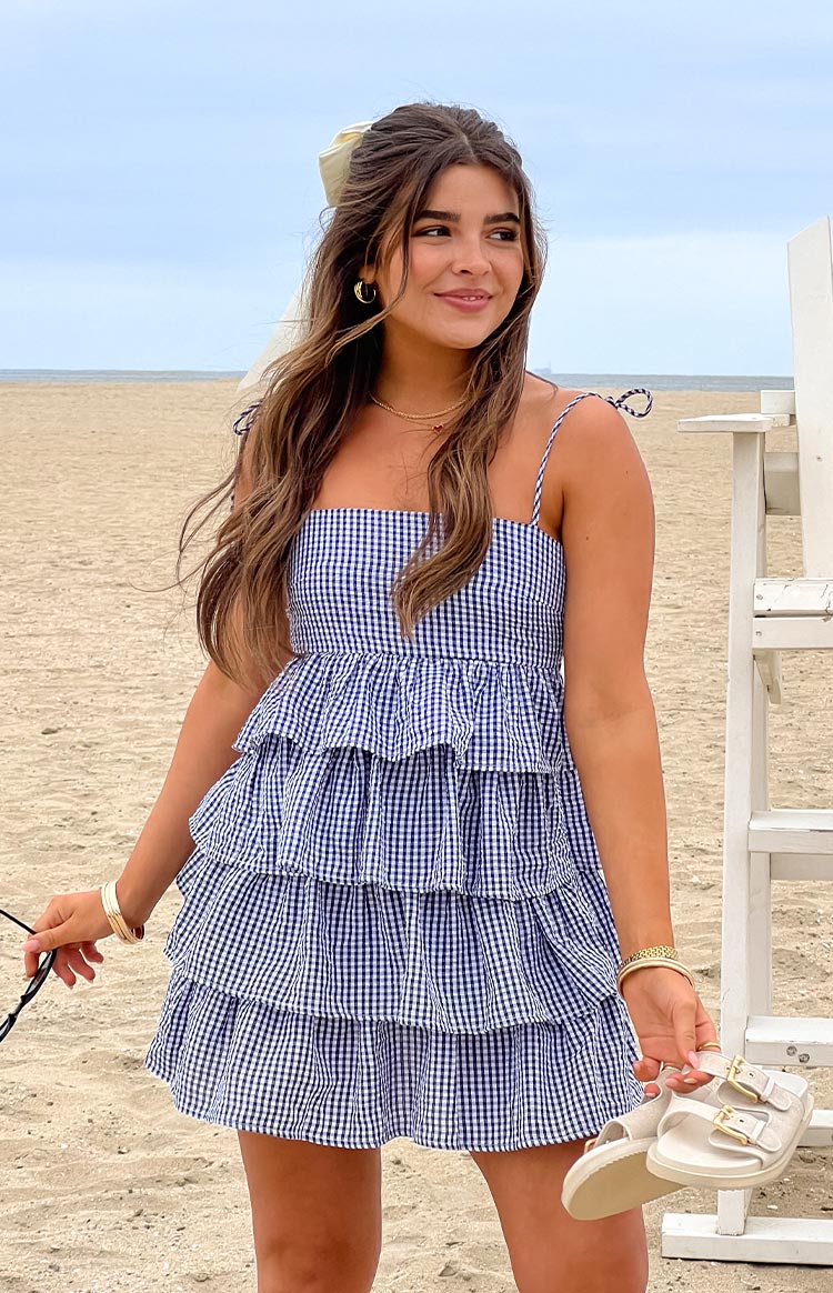 Front view of a navy gingham mini dress with a square neckline, tie shoulder straps, and a tiered ruffle skirt