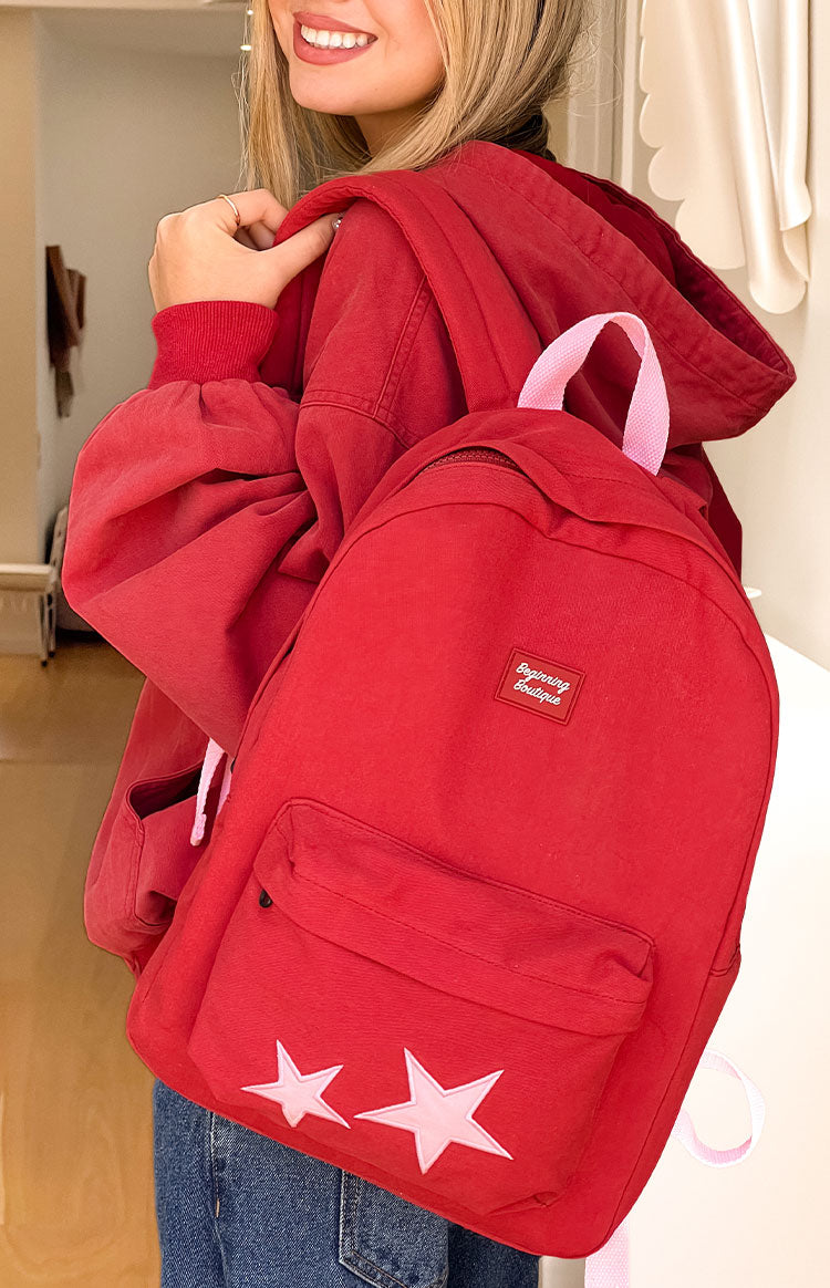 A front view of a red backpack with a zippered front pocket featuring two light pink star appliques.
