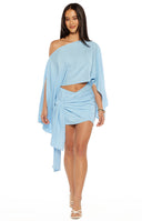 A lifestyle shot of our model wearing a light blue mini dress made from a textured linen-like fabric. The top is a cropped one-shoulder design with oversized draped sleeves that create a cape-like effect. The  mini skirt features an asymmetrical wrap front with a large decorative fabric knot at the hip and long hanging ties. These are connected in one side of waist part.